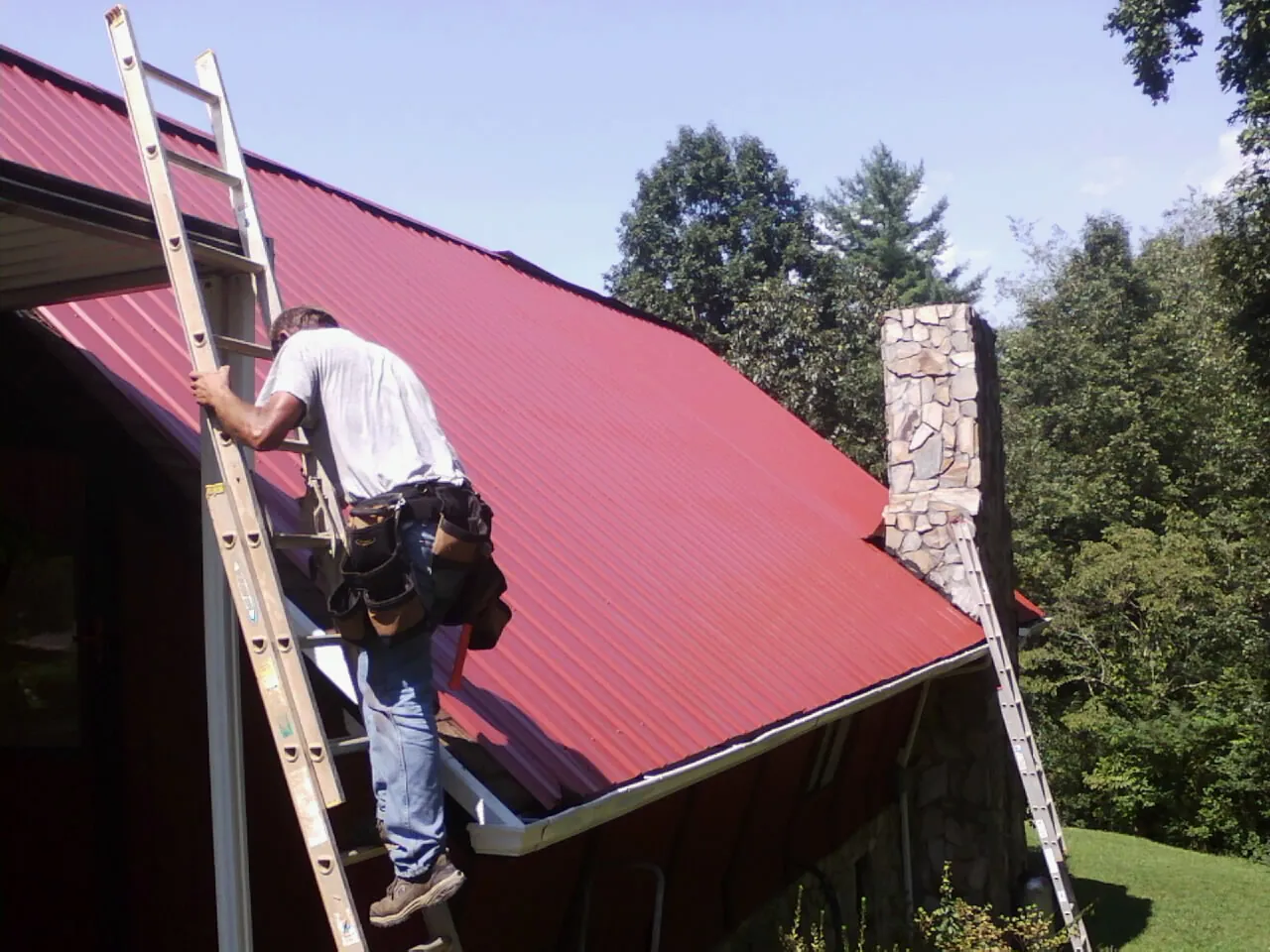 Roofer on a ladder beside a red metal roof and stone chimney — Soffit & Fascia Repair by Roofing Brothers in Pierson